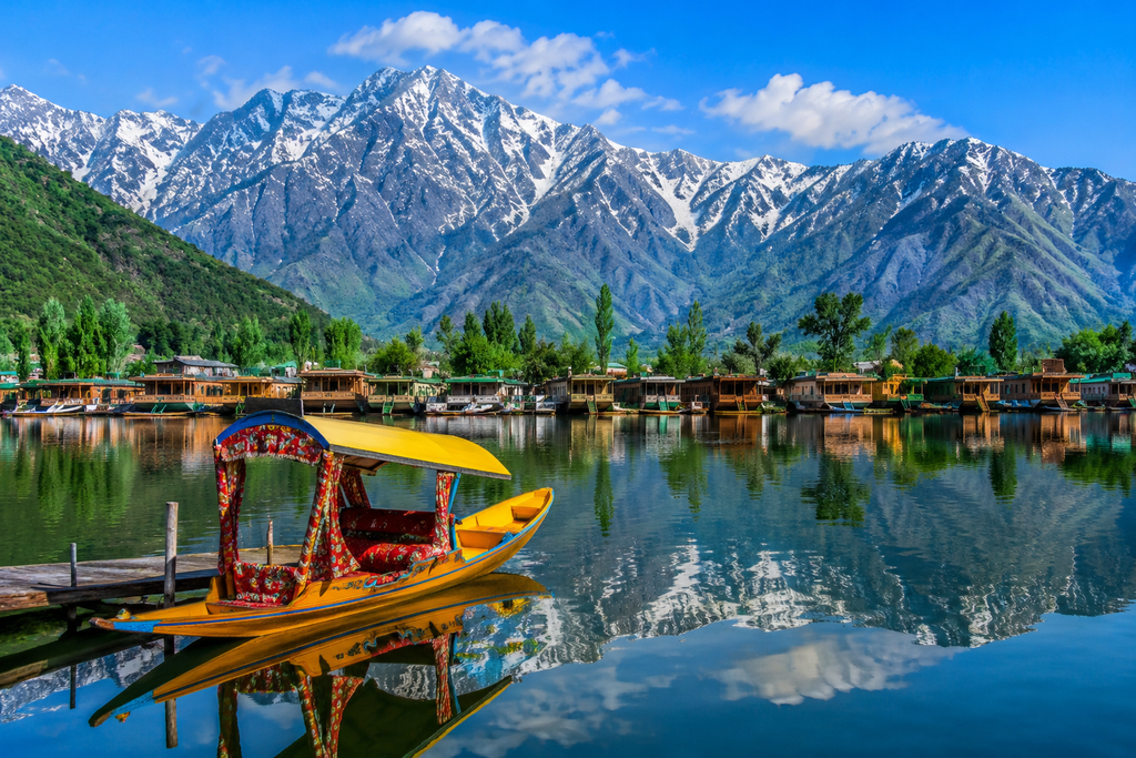 A colorful Kashmiri shikara boat on a calm lake, reflecting snow-capped Himalayan mountains, green trees, blue skies, and traditional houseboats along the shore.